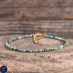 May include: A delicate turquoise beaded bracelet with a gold-colored clasp. The bracelet is made of small, round beads in shades of turquoise, green, and gray. The bracelet is displayed on a textured, brown surface. The text "@LND BANDS" is visible.