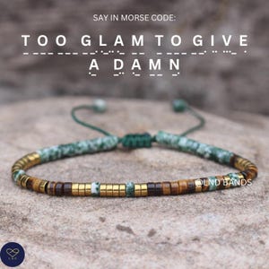 Peut inclure: Bracelet de perles vertes, dorées et marron. Le texte "TOO GLAM TO GIVE A DAMN" est écrit en code Morse. Le nom de la marque "OLND BANDS" est visible. Le bracelet est noué avec un cordon vert.