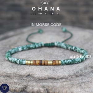 May include: A green and white beaded bracelet with gold and brown beads spelling "OHANA" in Morse code. The bracelet has a green adjustable cord. Text at the top says "SAY OHANA" and "IN MORSE CODE".