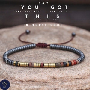 Puede incluir: Un brazalete de cuentas con cierre de cordón marrón. Las cuentas son negras, doradas y marrones. El brazalete tiene el texto "YOU GOT THIS" en código Morse.
