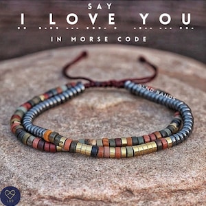 May include: A beaded bracelet featuring a dark grey, brown, gold, and red colour scheme. The bracelet is made of small, round beads and has a simple, adjustable clasp. The bracelet is on a brown, textured surface. The text "SAY I LOVE YOU IN MORSE CODE" is above the bracelet. The text "@LND BANDS" is on the right side of the bracelet.