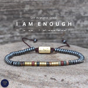 Op de afbeelding: Een kralenarmband met donkergrijze, gouden en roodbruine cilindrische kralen aan een donker koord. Een centrale goudkleurige rechthoekige kraal is gegraveerd met LND. De tekst erboven luidt SAY IN MORSE CODE: I AM ENOUGH met morsecode stippen en streepjes.