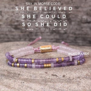 以下が含まれることがあります： 紫とゴールドのアクセントが付いた2つのビーズブレスレット。ブレスレットは重ねられています。「SAY IN MORSE CODE: SHE BELIEVED SHE COULD SO SHE DID」というテキストが画像に表示されています。