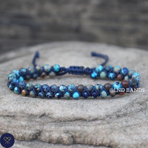May include: A blue beaded bracelet with a macrame design. The bracelet features a mix of blue, turquoise, and brown beads, strung on a dark blue cord. The bracelet is displayed on a gray stone surface.
