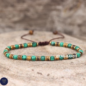 May include: A beaded bracelet featuring alternating green, brown, and gold-colored beads. The bracelet is strung on a brown cord and has an adjustable sliding knot closure. The bracelet is displayed on a textured, neutral-toned surface.