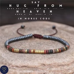 May include: A beaded bracelet with a dark grey, silver, and gold colour scheme. The bracelet has a sliding knot closure. The bracelet is made of small, round beads. The beads are arranged in a pattern of dark grey, silver, and gold. The bracelet is on a brown, textured surface. The text "SAY FROM HEAVEN IN MORSE CODE" is above the bracelet.