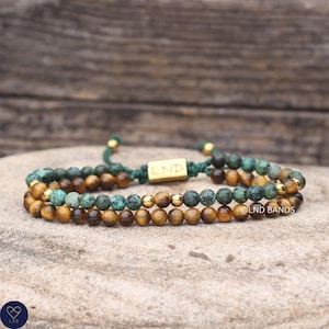 May include: A double-strand beaded bracelet featuring alternating round tiger eye and turquoise-coloured stone beads, accented with small gold-toned metal spheres. The bracelet includes a central gold-toned rectangular charm engraved with LND.