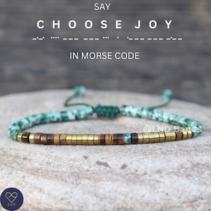 May include: A green and white beaded bracelet with gold and brown beads spelling out "CHOOSE JOY" in Morse code. The bracelet is tied with a green cord.