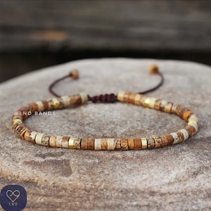 May include: A brown, white, and gold beaded bracelet with a brown cord closure. The bracelet has the text "@LND_BANDS" on it.