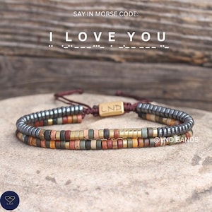 Op de afbeelding: Twee armbanden met kralen met een verscheidenheid aan aardetinten. De bovenste armband heeft donkergrijze kralen aan de uiteinden. De tekst "I LOVE YOU" is in morsecode boven de armbanden geschreven.