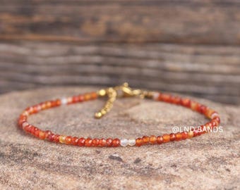 May include: A delicate bracelet with small, faceted orange and white beads. The bracelet has a gold-coloured clasp and chain. The text "@LND BANDS" is visible on the right. The bracelet is displayed on a textured, neutral-toned surface. A fine piece of jewellery.