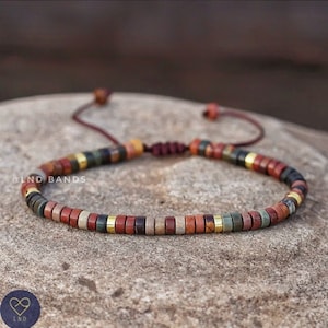 Puede incluir: Un brazalete de cuentas multicolores con cierre de cordón rojo. Las cuentas son una mezcla de marrón, rojo, verde y dorado. El brazalete está sobre una superficie rugosa y marrón. El texto "@LND_BANDS" es visible en la imagen.