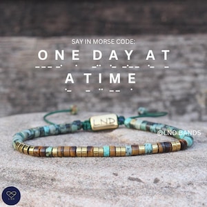 May include: A beaded bracelet featuring turquoise, brown, and gold-toned cylindrical beads on a green cord. A central gold-toned rectangular bead is engraved with LND. Text above reads: SAY IN MORSE CODE: ONE DAY AT A TIME with Morse code dots and dashes.