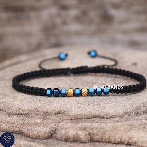 Può includere: Braccialetto in macramè nero con perline blu e oro. Il braccialetto ha una chiusura a nodo scorrevole e piccole perline blu. Il testo "@LND BANDS" è visibile sul lato destro dell'immagine.