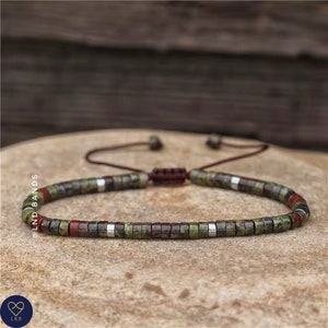 Puede incluir: Un brazalete de cuentas verdes, rojas y plateadas con cierre de cordón marrón. El brazalete está sobre una superficie marrón texturizada.