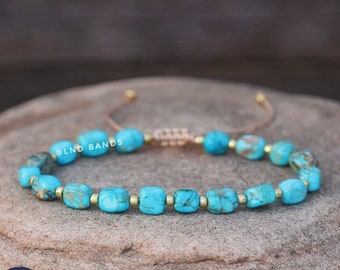 May include: A turquoise blue gemstone bracelet with gold beads and a brown cord. The bracelet has a sliding knot for adjusting the size. The bracelet is on a rough, textured surface.
