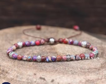 May include: A beaded bracelet with a mix of red, purple, and tan stones. The bracelet has silver-colored metal accents and a brown adjustable cord. The text "@LNDBANDS" is visible on the bracelet.