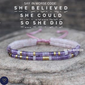 May include: A double-strand bracelet with purple and gold beads, and a pink cord. The text "SHE BELIEVED SHE COULD SO SHE DID" is above the bracelet in Morse code. The brand name "@LND BANDS" is also visible.
