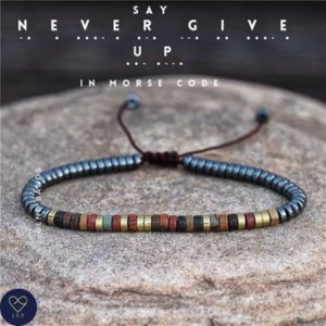 Könnte beinhalten: Ein Armband mit Perlen in einem dunklen Grau-, Schwarz- und Goldton. Das Armband hat einen verschiebbaren Knotenverschluss und trägt eine Botschaft in Morsecode, die "Gib niemals auf" lautet.
