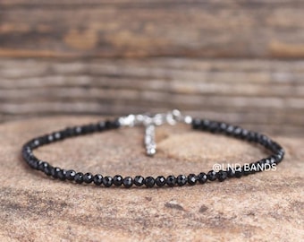 May include: A delicate black beaded bracelet with a silver-colored clasp. The bracelet is composed of small, round, black beads. The background is a textured, neutral-toned surface.