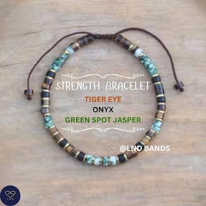 May include: A beaded bracelet with the text "STRENGTH BRACELET". The bracelet features tiger eye, onyx, and green spot jasper beads. The bracelet is brown, black, green, and gold. The brand name "@LND BANDS" is also visible.