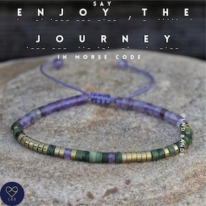 Puede incluir: Un brazalete de cuentas moradas y verdes con detalles dorados. El brazalete está atado con un cordón morado y dice "Say Enjoy the Journey, in Morse Code" sobre un fondo de piedra.