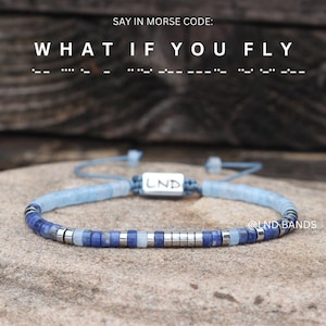 Könnte beinhalten: Ein blau-silbernes Perlenarmband mit dem Text "WHAT IF YOU FLY" in Morsecode. Das Armband hat blaue und silberne Perlen mit einem kleinen rechteckigen Silberanhänger mit der Aufschrift "LND". Das Armband wird auf einer Steinoberfläche präsentiert.