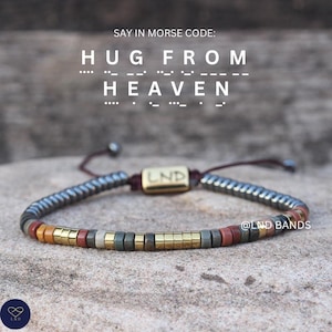 May include: A beaded bracelet with a mix of metallic, gold, and colorful beads. The bracelet has a dark red cord and a gold-colored rectangular charm with the letters "LND". The text "SAY IN MORSE CODE: HUG FROM HEAVEN" is at the top.