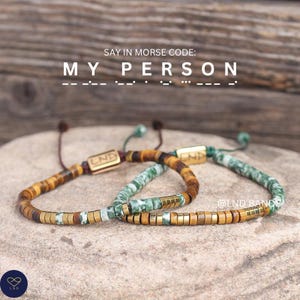 May include: Two beaded bracelets with the text "SAY IN MORSE CODE: MY PERSON". One bracelet features brown and gold beads, the other green and white beads. Both have gold-colored rectangular accents.