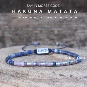 May include: A bracelet with blue, purple, and silver beads spelling "HAKUNA MATATA" in Morse code. The bracelet has the brand name "LND" on a silver bar. The text "SAY IN MORSE CODE" is at the top. The bracelet is on a stone surface.