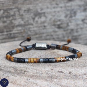 Könnte beinhalten: Ein Armband mit schwarzen, silbernen und braunen Perlen. Das Armband hat eine zentrale silberne rechteckige Perle mit den Buchstaben "LND" und ist mit einer schwarzen Kordel verstellbar. Der Text "@LND BANDS" ist ebenfalls sichtbar.