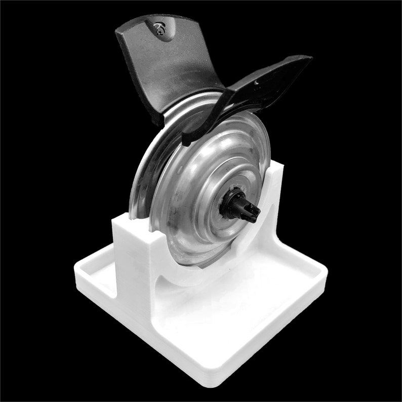 Pu&ograve; includere: Un supporto in plastica bianca con un oggetto rotondo di colore argento appoggiato su di esso. L'oggetto ha un coperchio in plastica nera con una piccola maniglia nera.