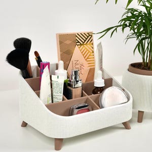May include: A white and tan desk organizer with multiple compartments containing various makeup brushes, skincare bottles, a compact, and an eyeshadow palette. The organizer features a ribbed exterior texture and stands on four small, rounded brown legs.