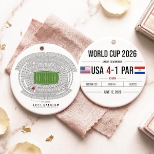 May include: Two round white ceramic ornaments. One features a stadium seating chart for SoFi Stadium in Inglewood, California, with a highlighted seat. The other displays World Cup 2026 text, USA 4-1 PAR score, flags, and seat details.
