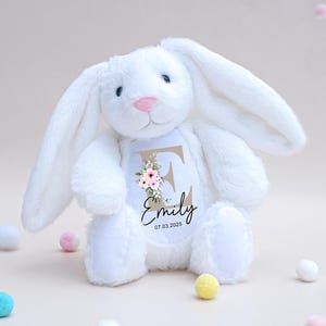 May include: A white plush bunny with long ears and a pink nose. The bunny has a personalized patch with the name "Emily", the initial "E", floral accents, and the date "07.03.2025". Decorative pom-poms in various colors are scattered around the bunny.