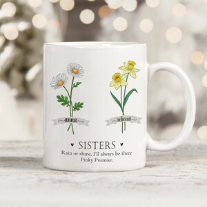 Puede incluir: Taza de cer&aacute;mica blanca con un dise&ntilde;o floral que presenta una margarita y un narciso. La taza tiene el texto "Anna" y "Rebecca" escrito en pancartas debajo de las flores. El texto "Sisters" est&aacute; escrito en forma de coraz&oacute;n con el texto "Rain or shine, I'll always be there Pinky Promise." debajo.