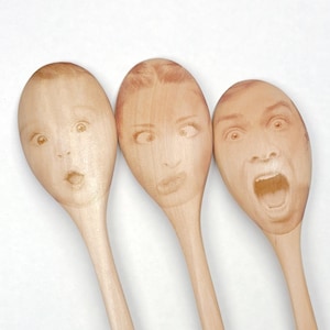 May include: Three wooden spoons with faces printed on the spoon heads. The faces have surprised, shocked, and screaming expressions. The spoons are made of light-colored wood and are arranged against a white background.