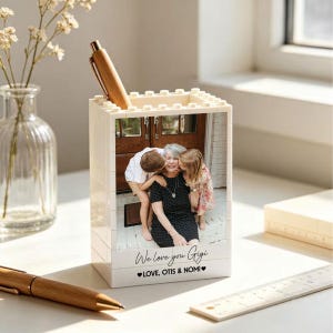 May include: A rectangular white plastic building block pen holder featuring a custom photo of an adult and two children. The base includes the printed text "We love you Gigi" followed by "LOVE, OTIS & NOMI" with two small black heart icons.