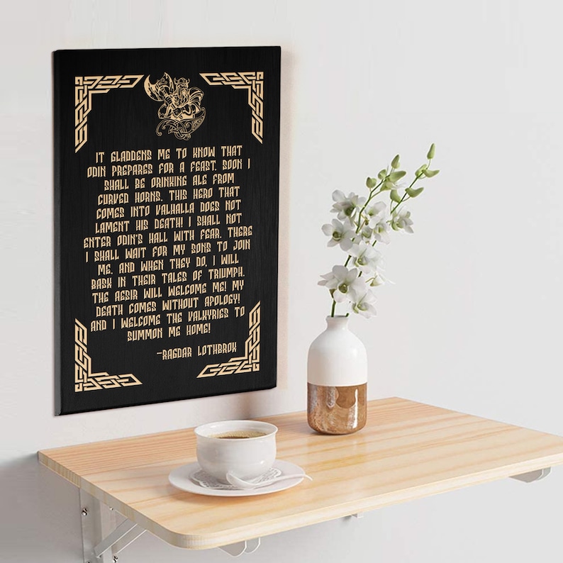 May include: A black wooden sign with gold lettering and a decorative border. The sign features a quote from the character Ragnar Lothbrok from the TV show Vikings. The quote reads: "It gladdens me to know that Odin prepares for a feast. Soon I shall be drinking ale from curved horns. This hero that comes into Valhalla does not lament his death! I shall not enter Odin's hall with fear. There I shall wait for my sons to join me. And when they do, I will bask in their tales of triumph. The Aesir will welcome me! My death comes without apology! And I welcome the Valkyries to summon me home!" -Ragnar Lothbrok