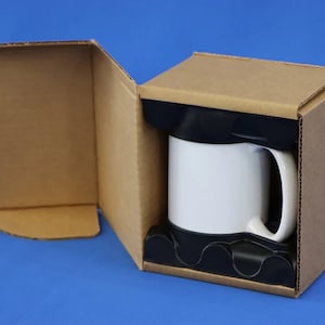 Puede incluir: Una taza de cer&aacute;mica blanca con asa negra est&aacute; empaquetada en una caja de cart&oacute;n marr&oacute;n con un inserto de espuma negra.