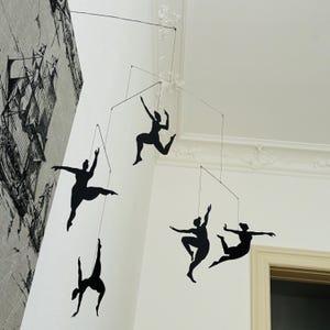 Puede incluir: Móvil moderno con siluetas negras de bailarines en diversas poses. Las figuras están suspendidas de finos alambres, creando una exhibición dinámica y artística. El móvil se encuentra contra una pared blanca con molduras decorativas.