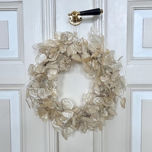 Può includere: Una ghirlanda rotonda beige fatta di foglie traslucide essiccate è appesa a una porta bianca. Sopra la ghirlanda c'è una maniglia in ottone e nera. La porta ha pannelli e finiture bianche.