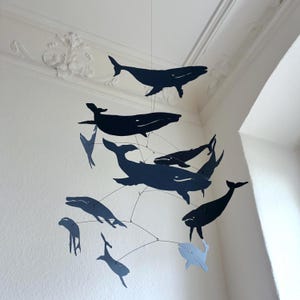 Op de afbeelding: Een decoratieve mobiel met walvisuitsparingen in blauwe en grijze tinten. De walvisvormen zijn opgehangen aan draden, wat een dynamische weergave creëert. Ideaal voor een kinderkamer, voor een speelse touch.