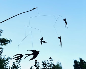 Könnte beinhalten: Ein schwarzes Metall-Mobile mit Vogel- und abstrakten menschlichen Silhouetten, die an dünnen Drähten hängen. Das Mobile hängt vor einem klaren, hellblauen Himmel, mit Ästen im unteren Bereich.
