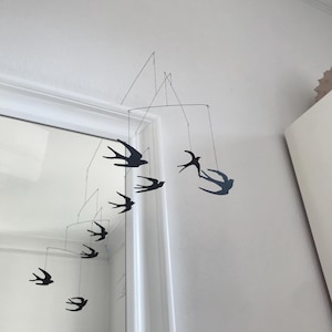 Può includere: Mobile in metallo nero con sagome di rondini sospese a fili sottili. Gli uccelli sono in varie posizioni, creando un elemento decorativo dinamico e arioso. Il mobile è appeso a una parete bianca.