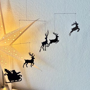 Può includere: Un mobile decorativo natalizio con sagome nere di renne e la slitta di Babbo Natale sospese con fili sottili. Il tutto è posto davanti a un muro bianco e una lampada a forma di stella.