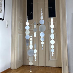 Puede incluir: Un móvil moderno con múltiples hilos de discos plateados, circulares y ovalados. Los discos varían en tamaño y están suspendidos de finos alambres, creando una pieza decorativa y dinámica. El móvil cuelga frente a una puerta clara.