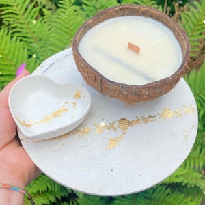 May include: A white heart-shaped dish with gold flecks sits on a white round tray with gold flecks. A coconut shell candle with a wooden wick sits on the tray.