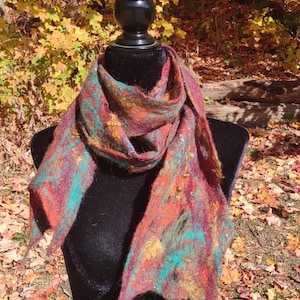 May include: A colorful scarf with shades of red, orange, green, and brown. The scarf is made of wool and has a fuzzy texture. It is displayed on a black mannequin against a backdrop of autumn leaves.