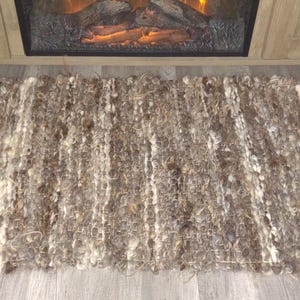 May include: A rectangular, hand-woven rug with a shaggy texture. The rug features a mix of brown and cream-colored fibers, creating a natural, earthy aesthetic. The rug is placed in front of a fireplace.